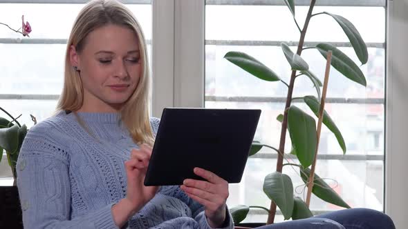 A Young Beautiful Woman Sits on a Chair By a Window and Works on a Tablet with a Smile