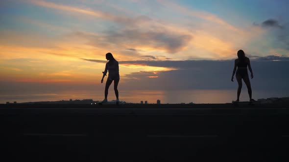 Silhouette of Two Girls Riding Skateboards on the Background of the Ocean and the Sunset Sky