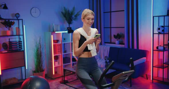 Woman in Sportswear Reading News on Smartphone while Exercising on Stationary Bike at Home