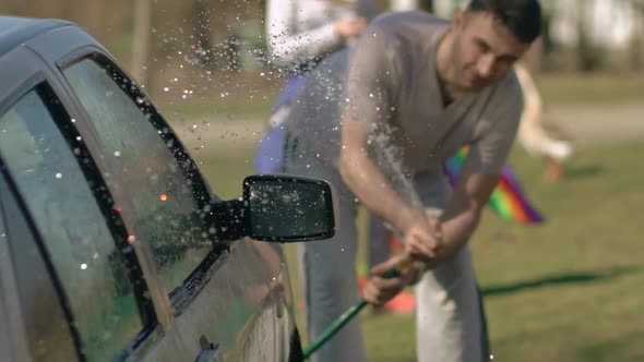 An Adult Man Washes His Car By Spraying Water From a Hose