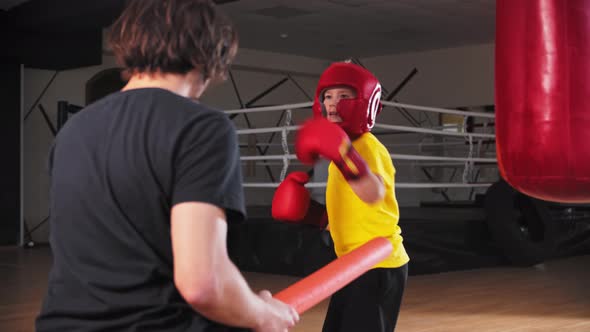 Little Boy in Soft Protective Helmet Doing Boxing with a Young Coach Hitting in the Long Soft Sticks