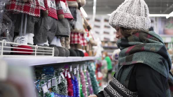 A Young Woman in a Black Down Jacket in a Supermarket Chooses Christmas Decorations