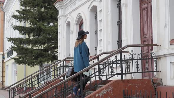 Young Redhaired Woman in a Blue Coat and Black Hat with a Suitcase on the Background of the Old City