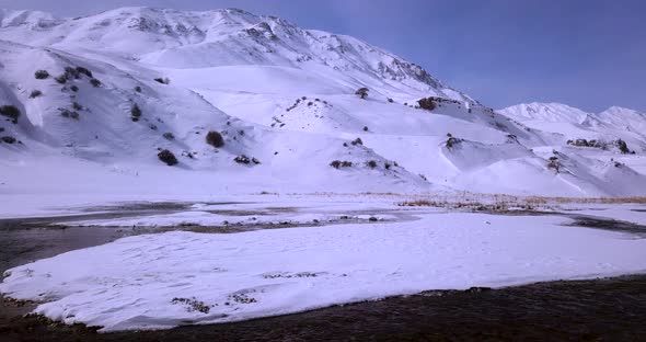 Fly Over A Frozen Icy River Stream Flow And White Snow Covered Mountain Hills Landscape In Backgroun