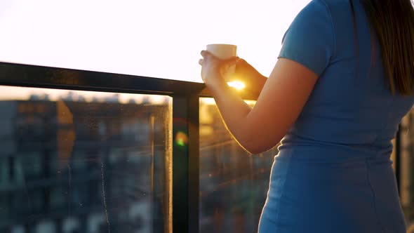 Woman with a Cup of Coffee Standing on the Balcony and Admire the Sunset