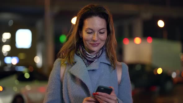 Portrait of a Smiling Blonde in a Light Coat and Scarf with a Phone in Hands Against the Background