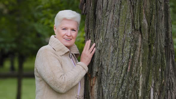 Happy Calm Mature Middle Aged Single Woman Looking at Camera Posing Near Large Tree Smiling Older