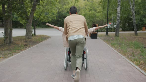 Caring Husband Running and Pushing Wheelchair Having Fun with Disabled Wife Outside in Autumn Park