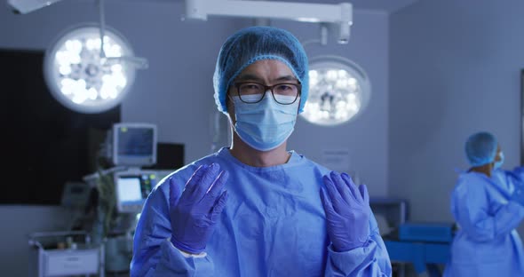 Portrait of asian male surgeon wearing face mask, gloves, cap and scrubs in operating theatre
