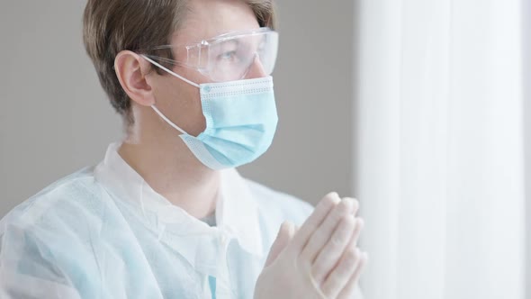 Portrait of Frustrated Young Male Doctor in Covid Face Mask Praying Standing at Window Indoors