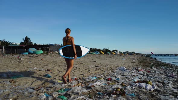 Athletic surfer girl with surfboard strolls on beach polluted with plastics