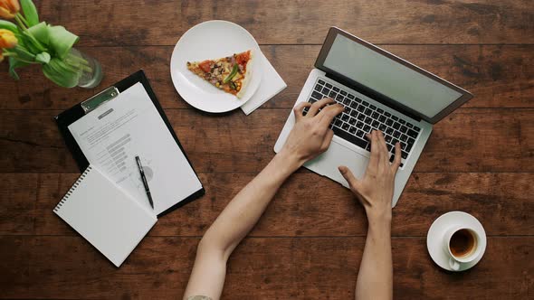 Man's Arms Typing on Laptop Wooden Table Pizza Slice Aside Cup of Coffee Office Papers Shot From