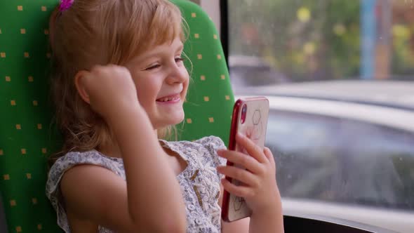 Smiling Child Girl in City Bus or Tram Transport Use Mobile Cell Phone Typing Browsing Say Wow Yes