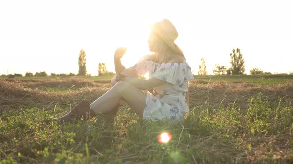 A Young Beautiful Woman in a Dress Sits in a Field Against Background of a Sunset