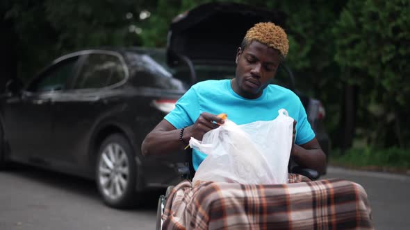 African American Young Man in Wheelchair with Groceries Outdoors with Blurred Black Car at