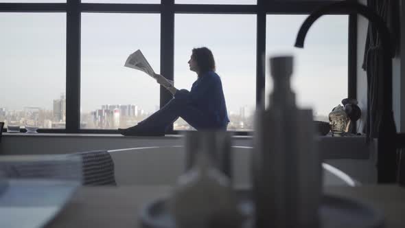 Serious Caucasian Businesswoman Sitting on Windowsill and Reading Newspaper. Side View Portrait of