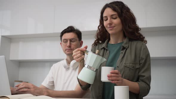 Caring Wife Pours Coffee for Her Husband While He is Working on a Laptop Sitting in the Kitchen