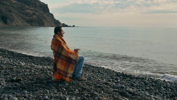 Slow Motion Young Man with a Beard and Mustache Sits in a Blanket and Coughs on a Deserted Seashore