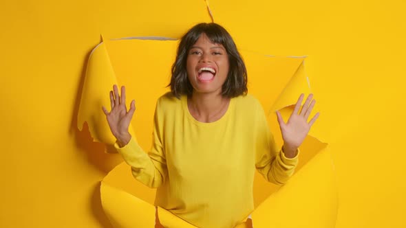Young Woman Exclaim with Joy Raises Hands in Ripped Hole Yellow Background