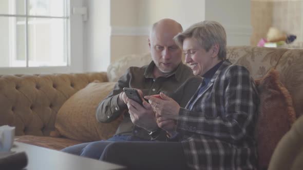 Senior Couple Sitting on the Big Sofa and Bald Man Showing Photos in the Mobile Phone for His Woman