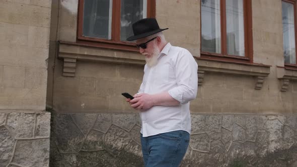 Old Man with Albinism Browsing on the Internet While Walking Next To Old House