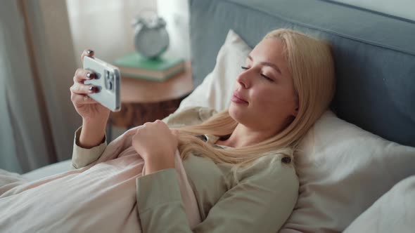 Positive blond woman hiding behind a blanket and looking at phone in bed