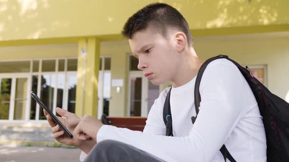 A Caucasian Teenage Boy Works on a Smartphone As He Sits in Front of School  Side Closeup