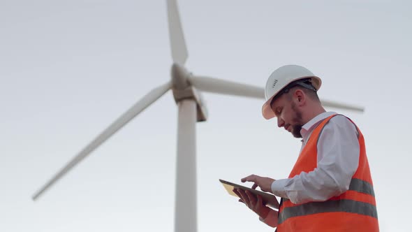 A Male Engineer in an Orange Vest and White Hard Hat Against the Background of a Windmill Looks and