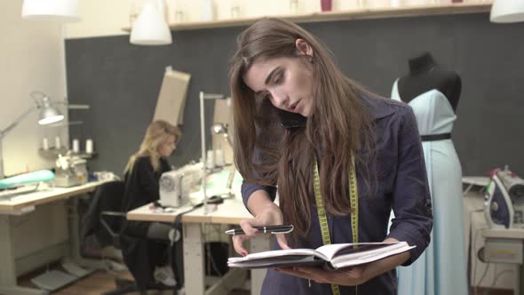 Busy Caucasian Brunette Young Female Standing in Sewing Classroom with Notebook Talking on Mobile