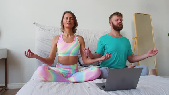 A Middleaged Couple Meditates in Front of a Laptop Monitor on a Bed Under the Guidance of an Online