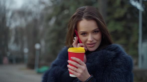 Attractive girl outdoors. Young woman in fur coat talking the phone while drinking coffee