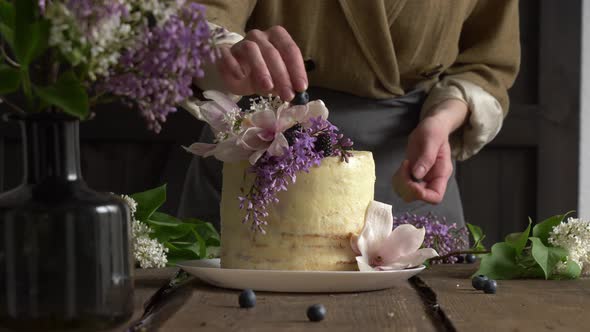 Pastry chef is decorated white wedding cake with lilac and magnolia flowers on a table