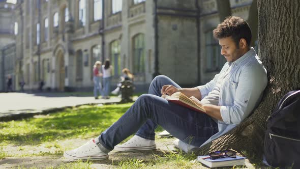 Multinational Guy Sitting Under Tree, Reading Book Looking Around, Leisure Time