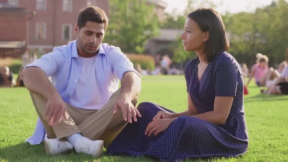 Happy Multiethnic Young Couple Sitting on Green Lawn in Park and Talking