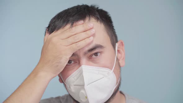 A man in a protective medical mask rubs his sore head with his palm and coughs into the mask.