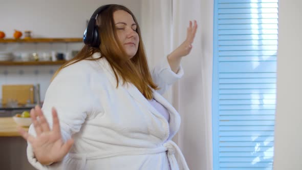 Cheerful Fat Woman Listening To Music in Headphones and Dancing Near Window at Home