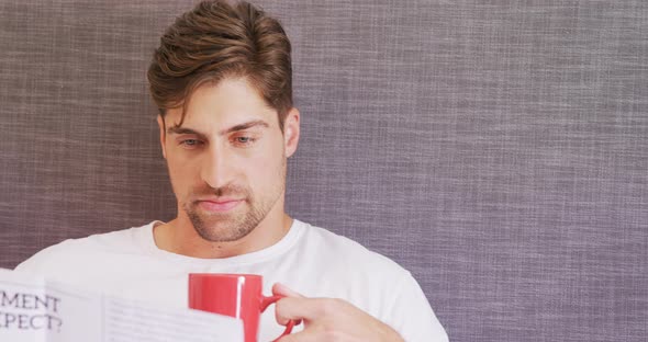 Young man sitting on bed having coffee while reading newspaper 4K 