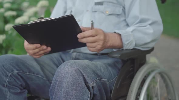 Unrecognizable Paraplegic Senior Man Signing Documents Outdoors. Old Caucasian Paralyzed Retiree