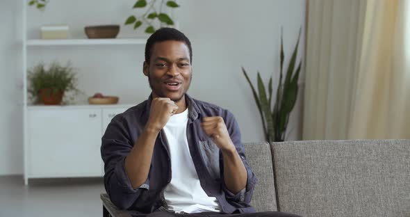 Young Enthusiastic African Guy Sitting at Home in Living Room on Couch Watching TV Online