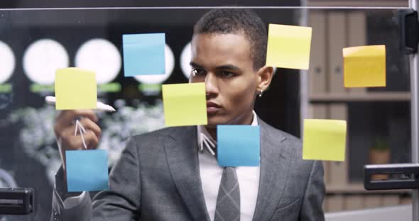 Young African Man in a Modern Office, Draws Arrows Infographic on the Glass Board with Sticky Notes