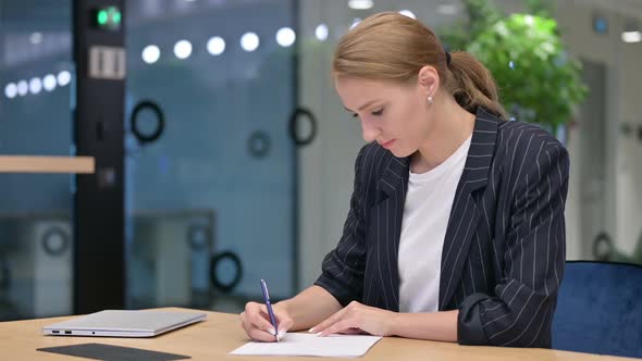 Serious Young Businesswoman Writing on Paper in Office