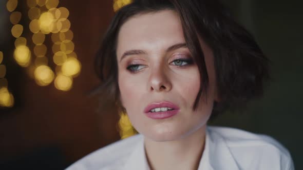 Thoughtful Woman with Makeup in White Shirt Looks at Camera with Cute Smile