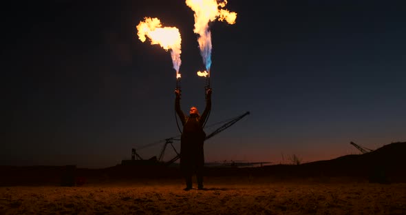 A Man in a Raincoat with Two Flamethrowers Lets Out a Fiery Flame Standing at Sunset on the Sand