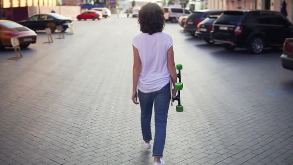 Back View of a Woman Walking in the Old City Street Holding Her Longboard in the Morning Parking in