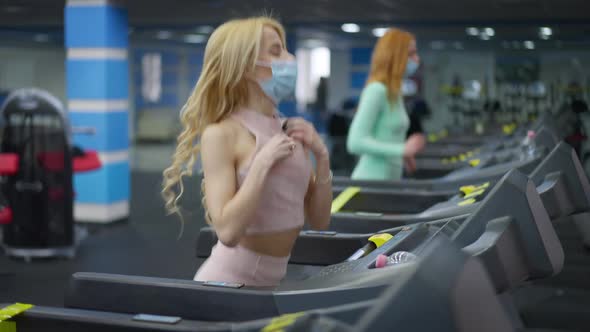 Side View Portrait of Blond Young Fit Woman in Coronavirus Face Mask Running on Treadmill As Blurred
