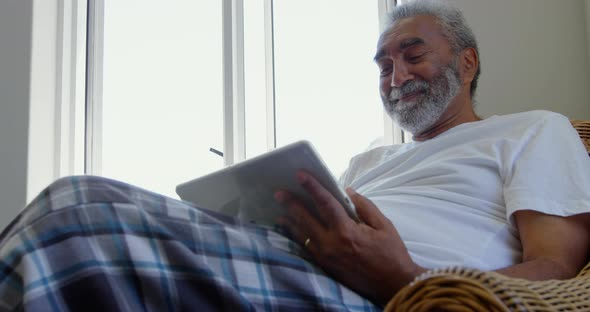 Front view of senior black man sitting on the couch and using digital tablet in comfortable home 4k