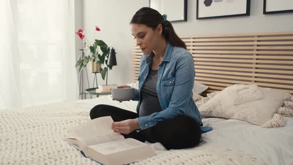 Caucasian woman in advanced pregnancy sitting on bed, eating salad and reading book.