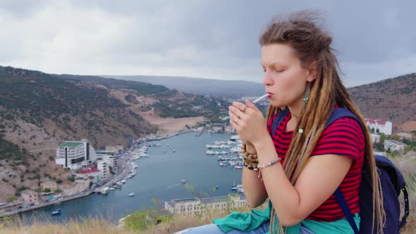 Girl Tourist with Dreadlocks Sits on Top of Hill Smokes