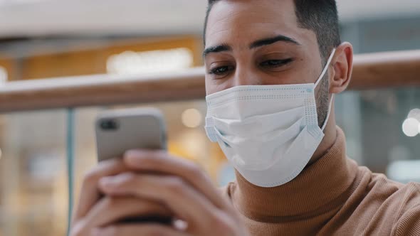 Closeup Young Happy Arab Guy in Medical Mask Holding Mobile Phone Smiling Surfing Web on Dating Site