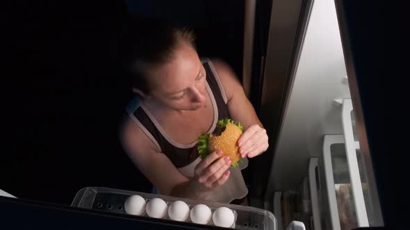 Woman Eating a Burger Near the Fridge